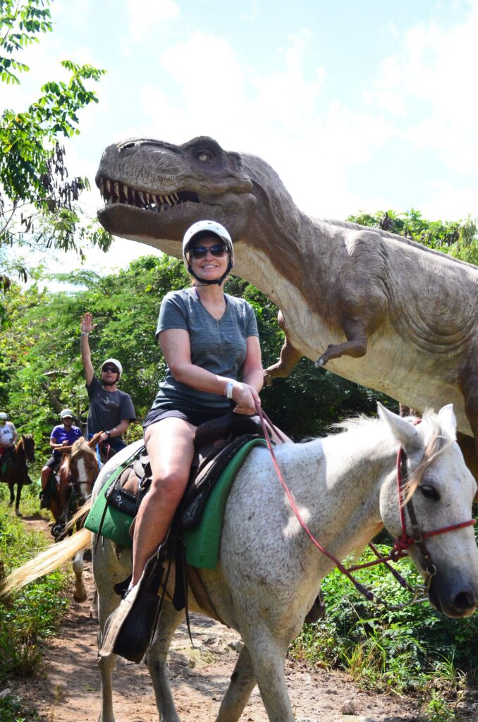 Smiling woman on horseback beside a life-size T-rex dinosaur statue on a jungle trail