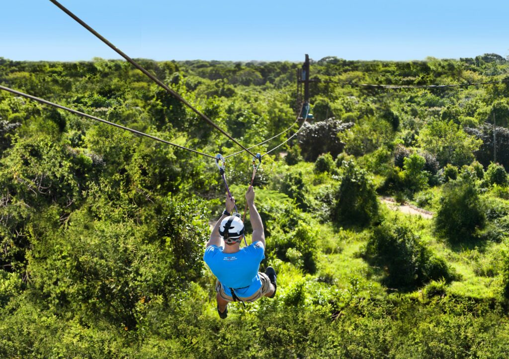 Man in blue shirt and helmet ziplining over vast green jungle canopy under blue sky