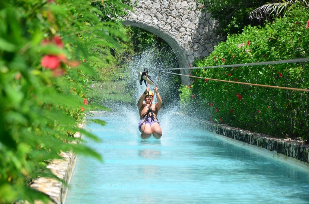 Woman ziplining down a turquoise water channel toward a stone bridge in a tropical garden