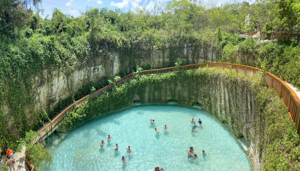 Tourists swimming in a turquoise natural cenote pool surrounded by cliffs and greenery
