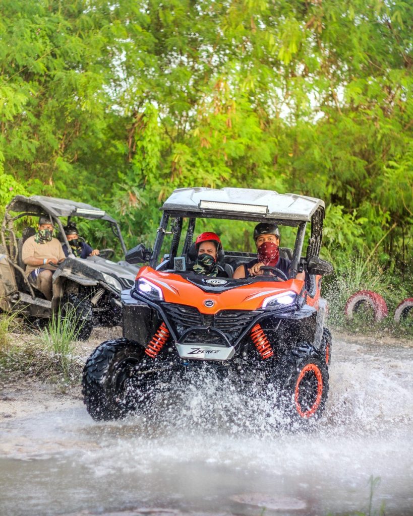 Orange CFMoto buggy splashing through water on a tree-lined off-road trail with riders in bandanas
