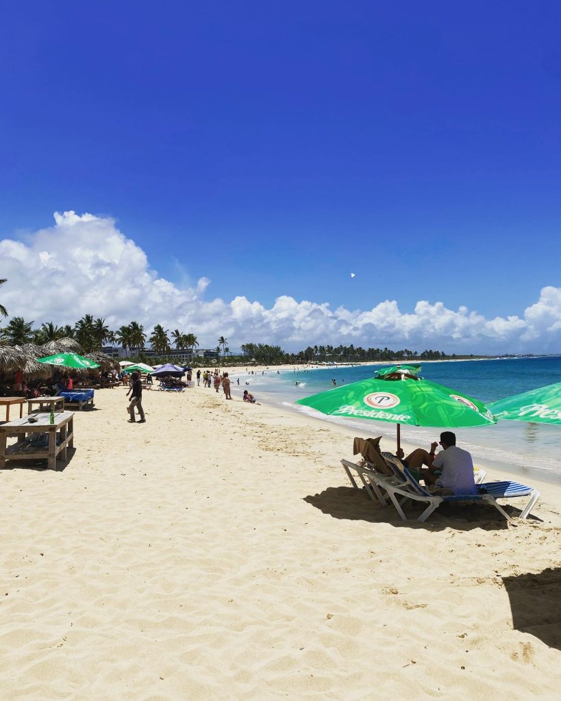 Sunny Caribbean beach with lounge chairs, green umbrellas, palm trees, and calm blue water