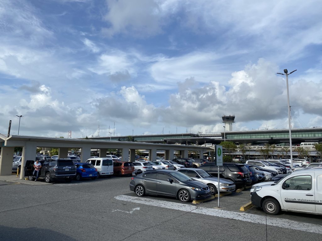 Airport parking lot with parked cars and a terminal building under a cloudy sky