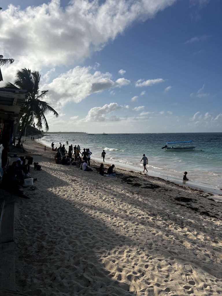 Playa al atardecer con personas, palmeras, un bote anclado y agua turquesa