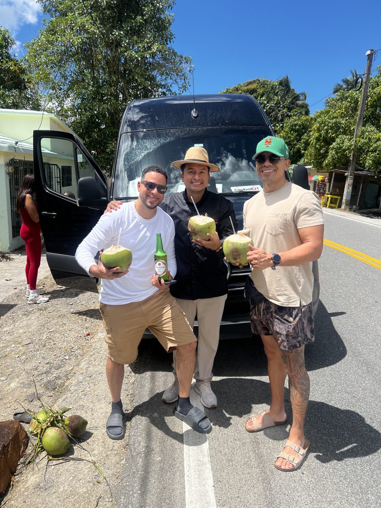 Three men holding fresh green coconuts beside a GlobalDT vehicle on a roadside stop