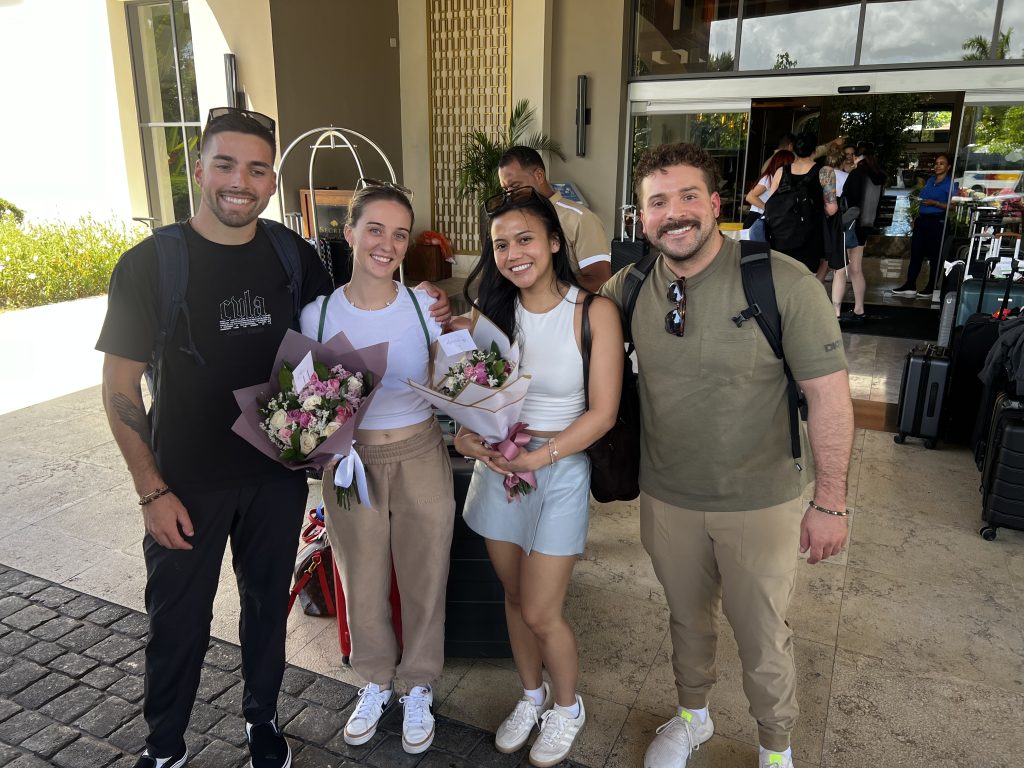 Two couples smiling at a resort lobby holding welcome flower bouquets with luggage